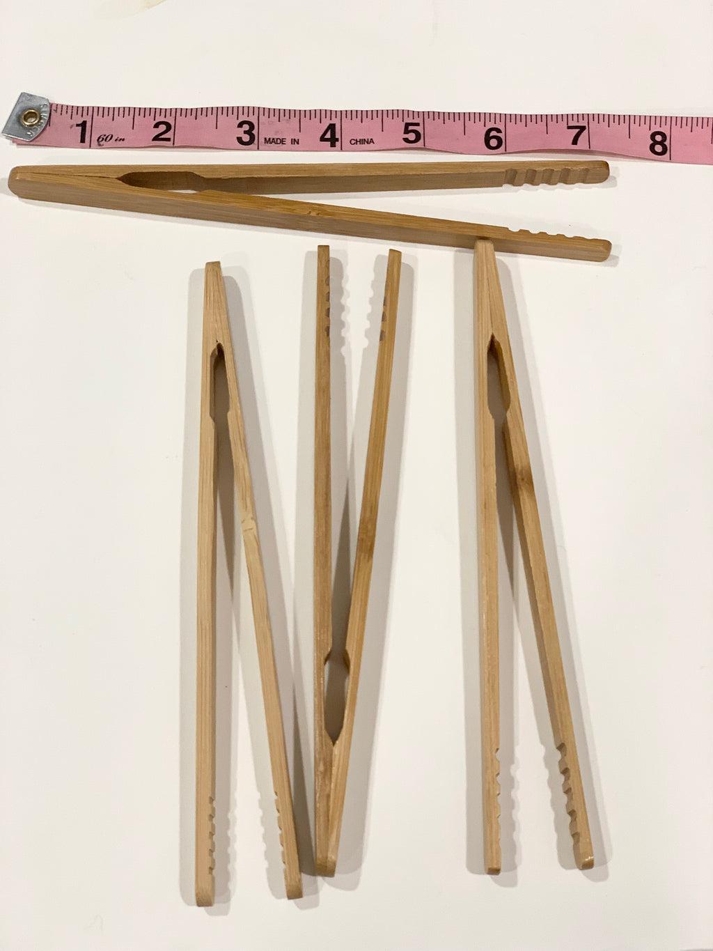 Set of five natural wooden sensory bin tongs with textured grip edges laid flat near pink measuring tape on white background