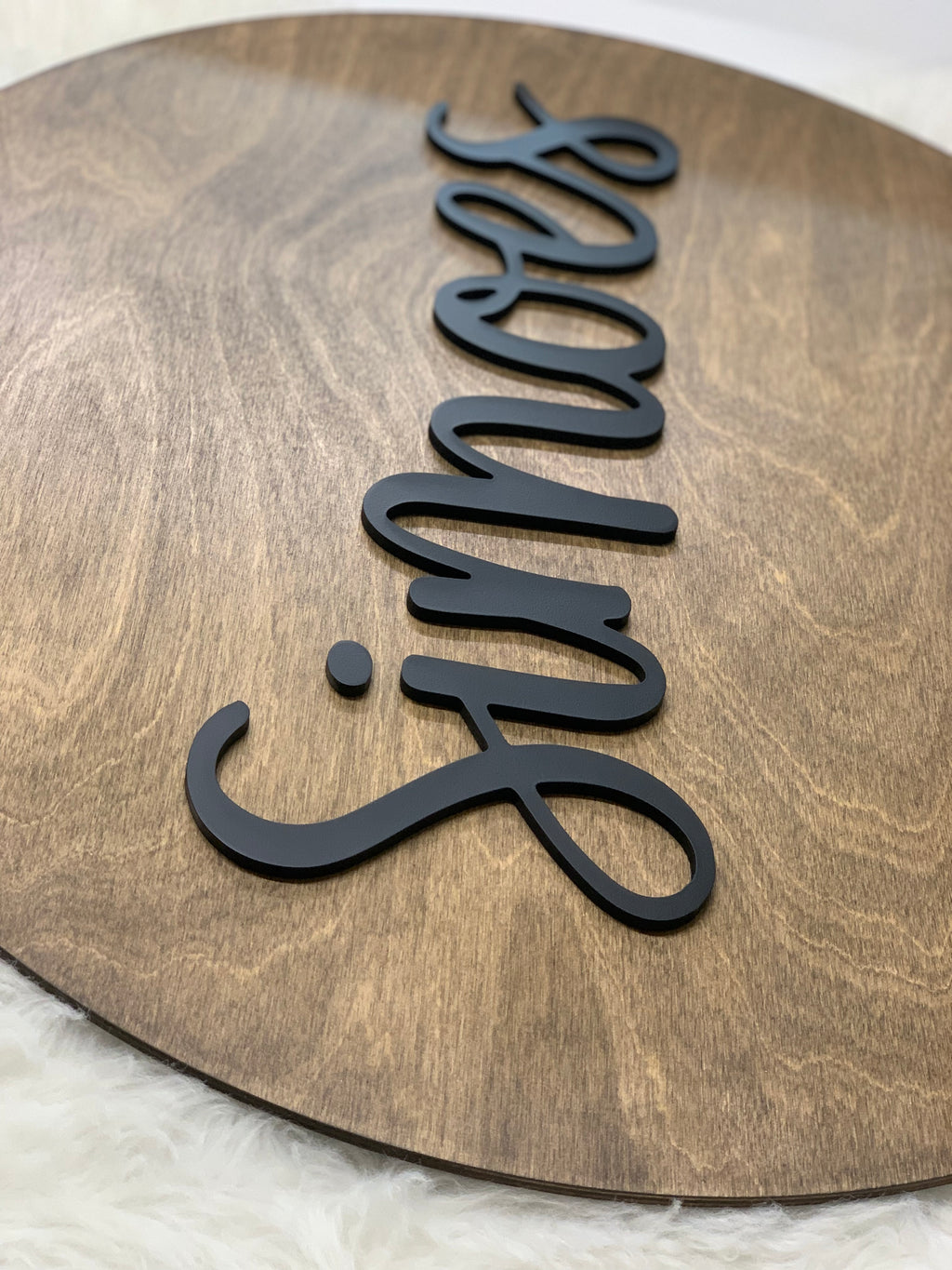 Round wooden name sign with black 3D cursive lettering on natural wood grain background