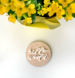 Round natural wooden disc with white cursive "hello world" lettering on white background with yellow flowers top left