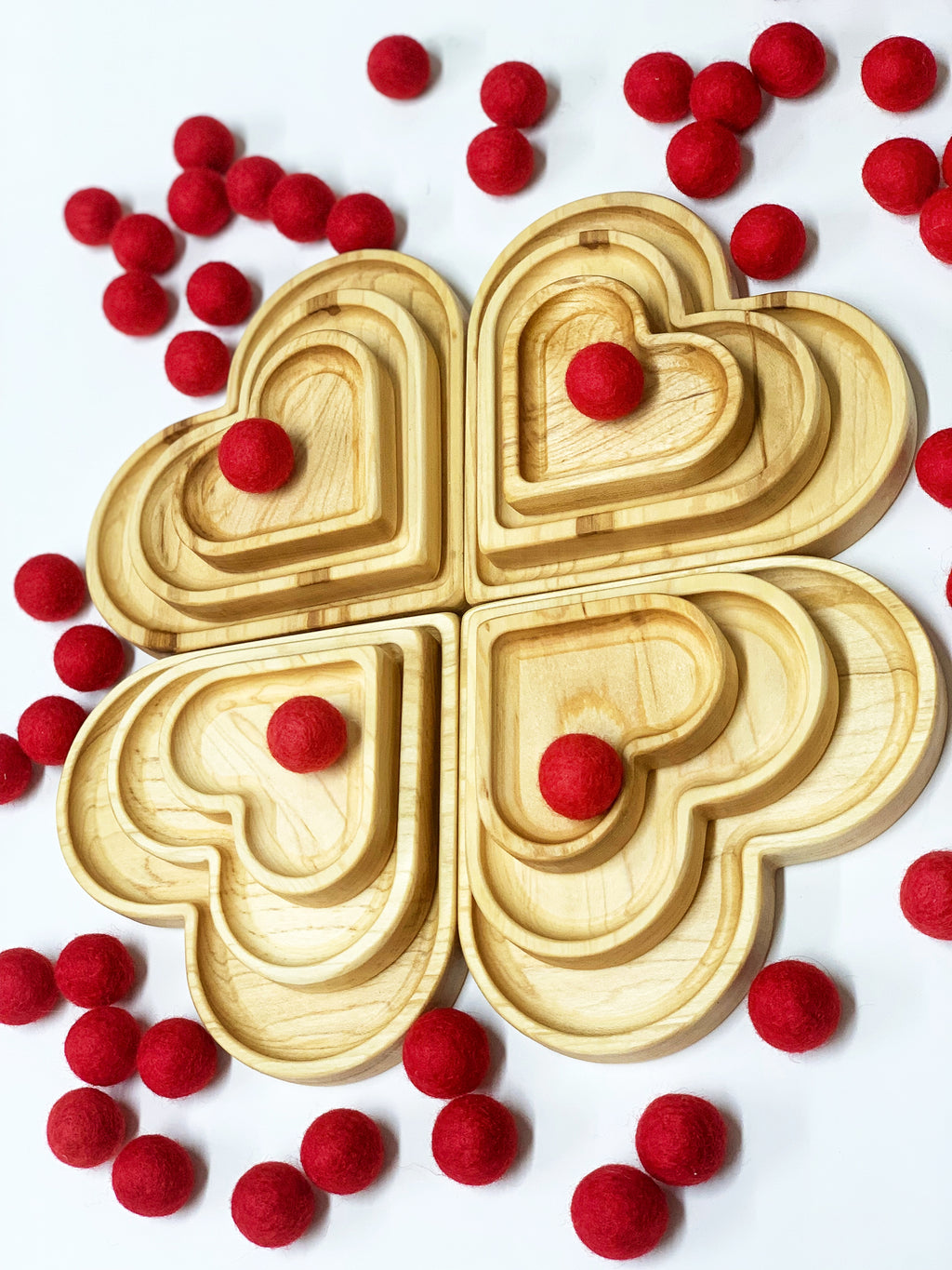 Natural wooden nesting heart plates arranged in clover shape with scattered red felt balls on white background