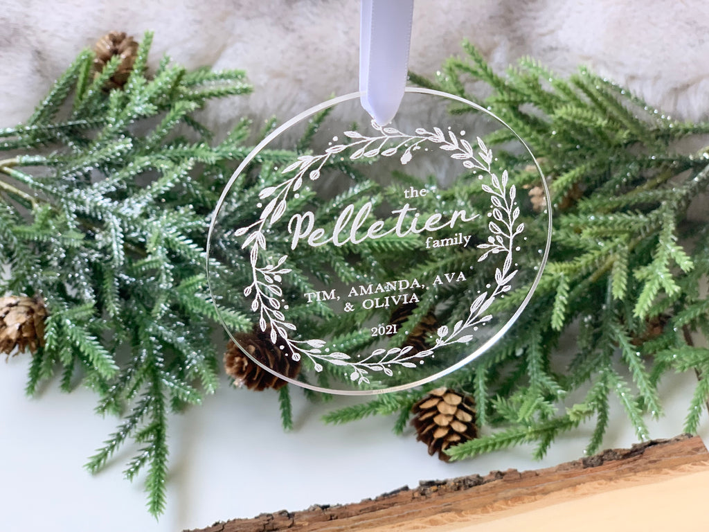 Clear acrylic Christmas ornament engraved with family name and decorative leaf wreath hanging with ribbon over pine branches