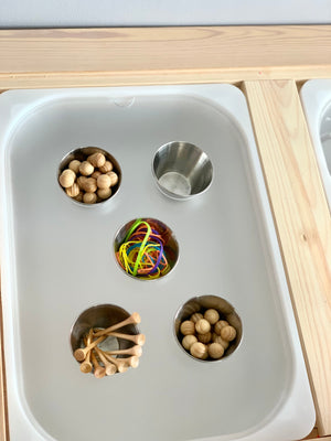 White acrylic insert with four metal cups holding wooden balls, pegs, and colorful rubber bands on light wood frame