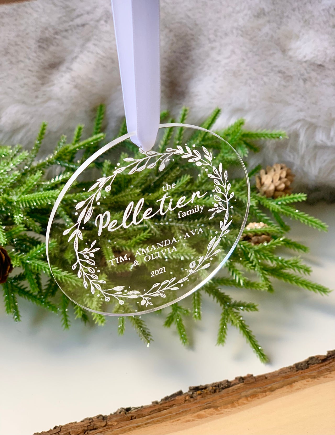 Clear round acrylic family Christmas ornament engraved with white script and floral wreath design hanging on white ribbon