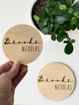 Round baby name announcement disc with cursive first name and capitalized middle name on natural maple wood background