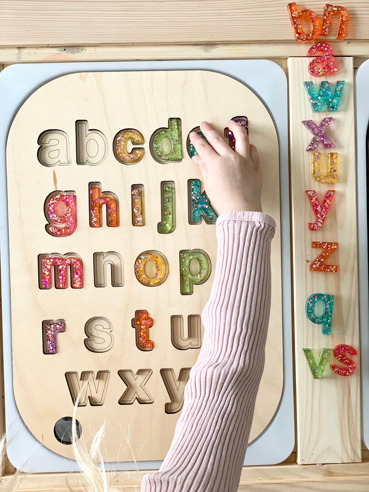 Child placing glitter resin lowercase letters into wooden alphabet puzzle with a drawstring bag and wooden board on table