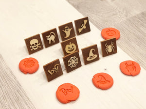 Set of small wooden Halloween play dough stamps with skull, cat, ghost, bat, pumpkin, spider, spider web, witch hat, and Boo designs on gray floor