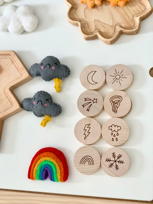Natural wooden 2-inch discs engraved with weather symbols arranged on white surface with felt cloud and rainbow toys