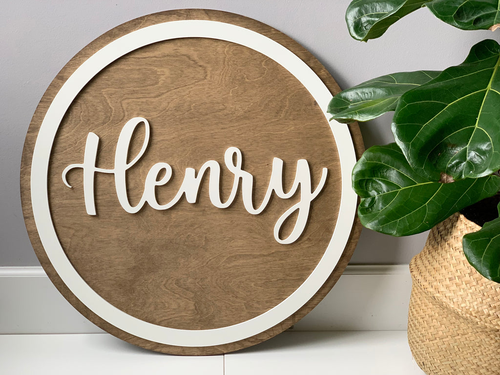 3D round wooden name sign with white cursive letters and white border on natural wood background next to potted green plant
