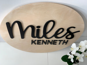 Oval natural wooden 3D name sign with black cursive and block letters spelling Miles Kenneth on white surface with white flowers