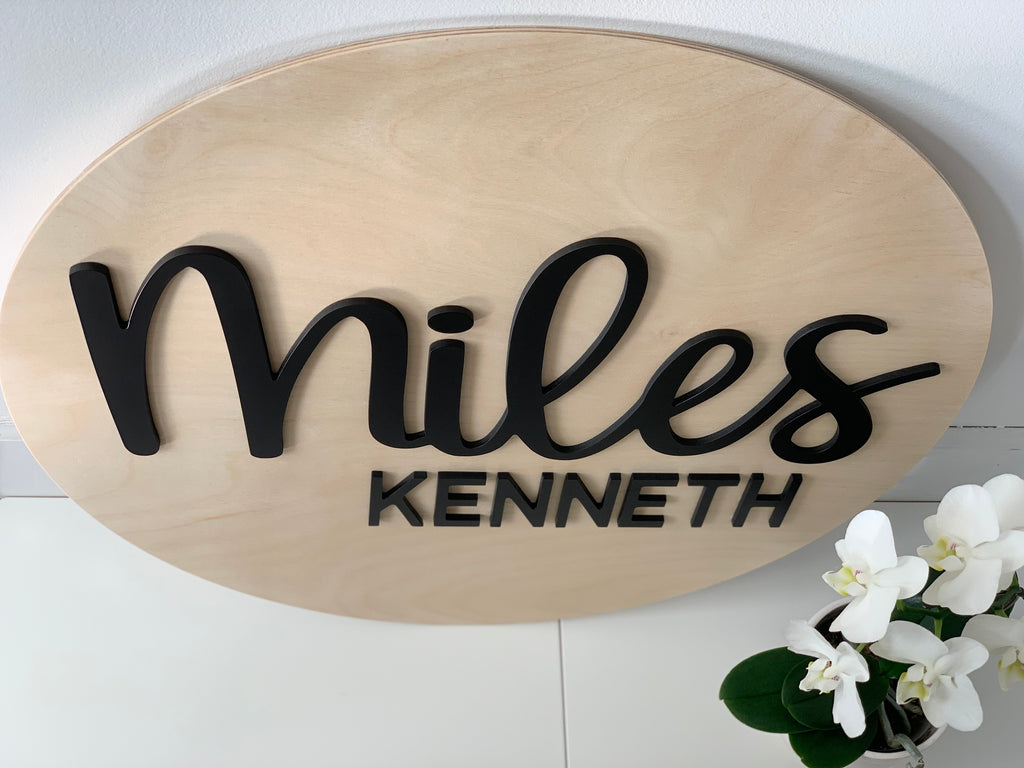 Oval natural wooden 3D name sign with black cursive and block letters spelling Miles Kenneth on white surface with white flowers