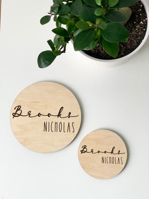 Two personalized round wooden baby name announcement discs with "Brooks Nicholas" engraved near green potted plant on white surface