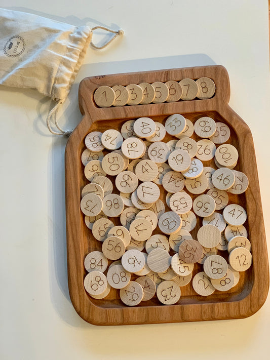 Mini wooden number discs 1 to 100 in jar-shaped tray with drawstring storage bag on white surface
