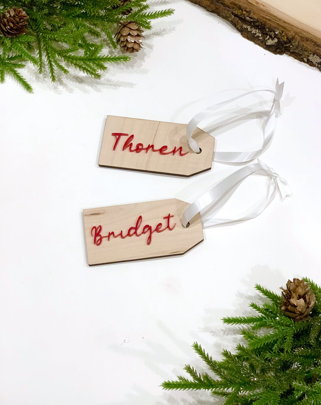 Custom wooden 2x4 inch gift tags with red cursive names and white ribbons on a white surface with pine and pinecones