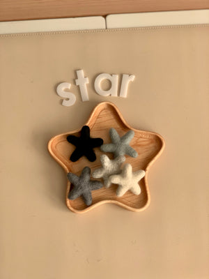 Small handmade neutral felt stars in black, gray, and white arranged inside wooden star-shaped tray on beige surface