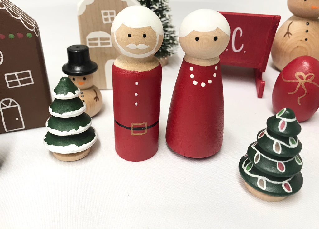 Hand-painted wooden Christmas peg dolls including Santa, Mrs Claus, decorated trees, and festive holiday figures on white surface