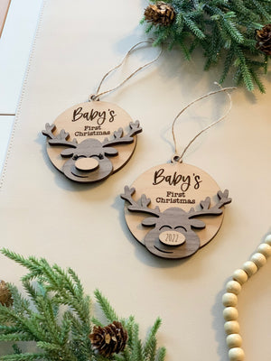 Pair of wooden "Baby's First Christmas" ornaments with moose designs and twine hangers on cream surface with greenery accents