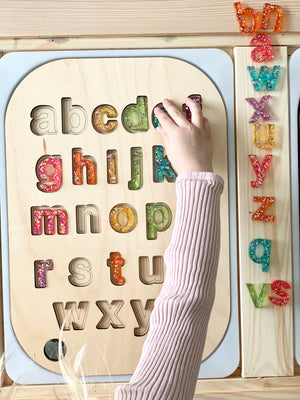 Wooden lowercase alphabet puzzle insert with colorful resin letters and child's hand placing letter on Flisat table top