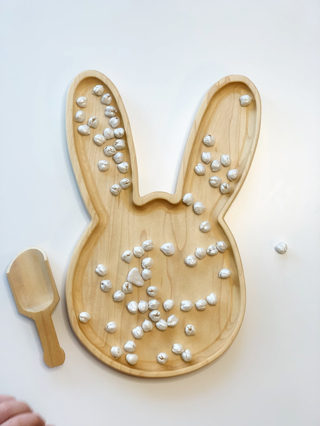 Large wooden bunny-shaped sensory tray with scattered small white textured beads and matching wooden scoop on white surface