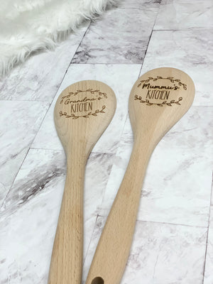 Two personalized wooden kitchen spoons engraved with "Grandma's Kitchen" and "Mummu's Kitchen" on light marble surface