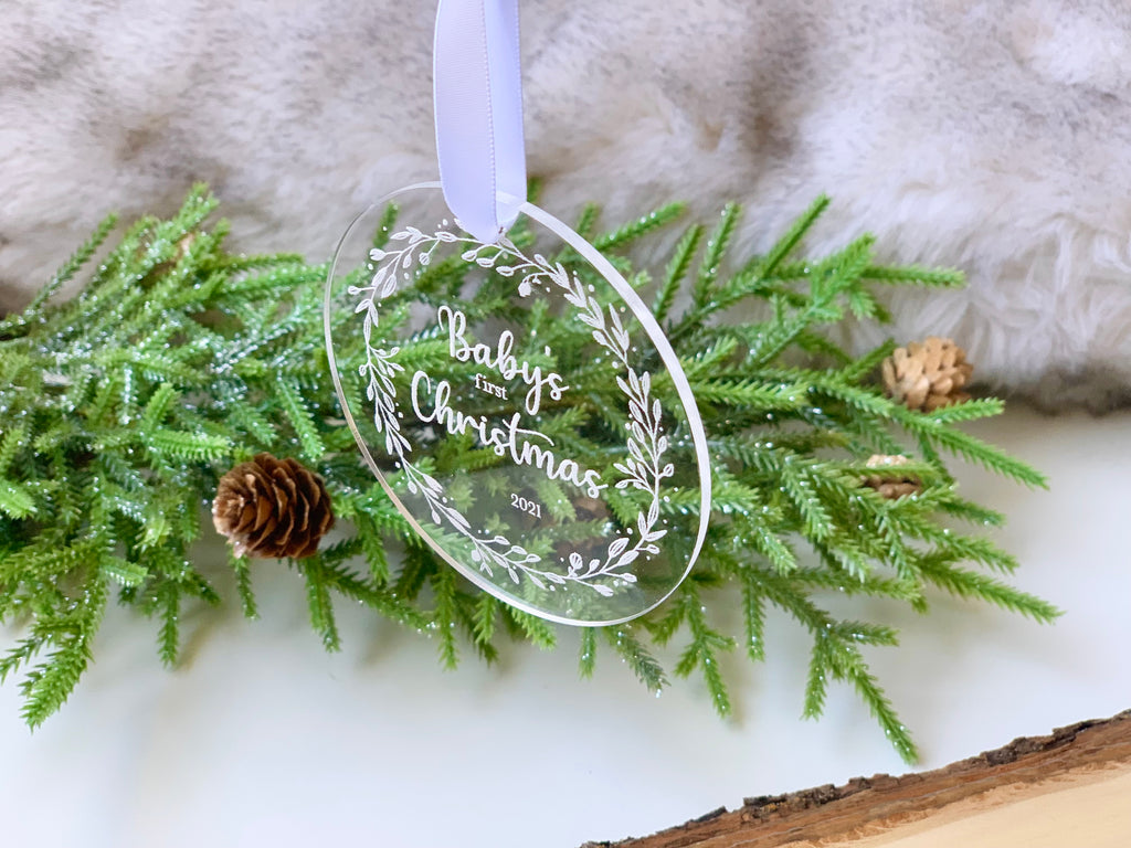 Clear acrylic round Christmas ornament engraved with "Baby's First Christmas 2021" and white ribbon on greenery background