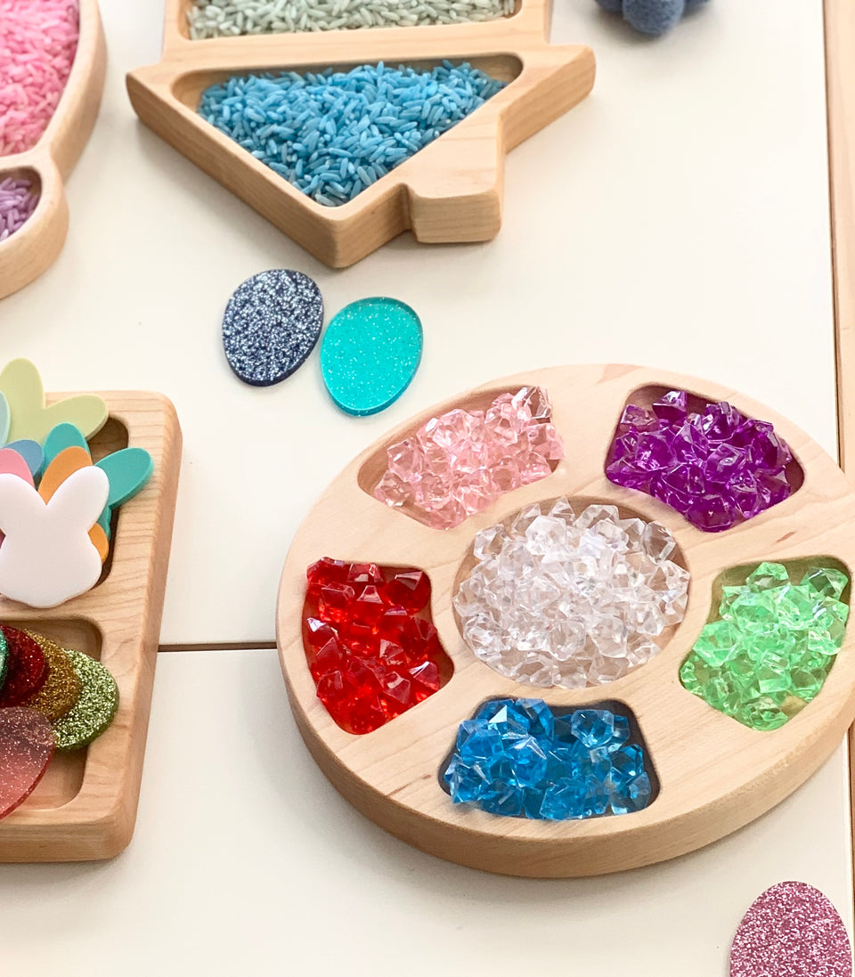 Round natural wooden tray holding assorted colorful acrylic treasure gems on white surface with craft materials nearby