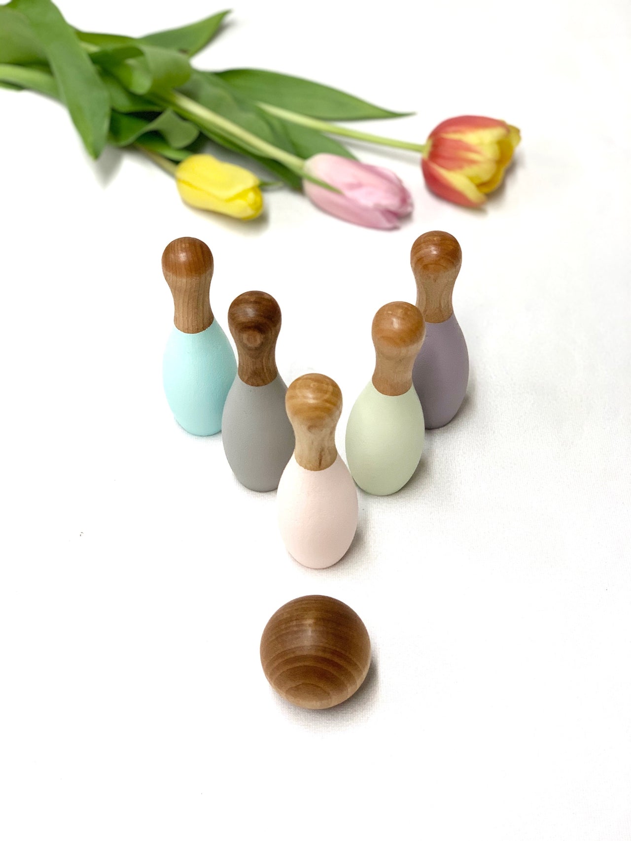 Pastel colored wooden bowling pins and ball for toddlers with tulips in the background on white surface