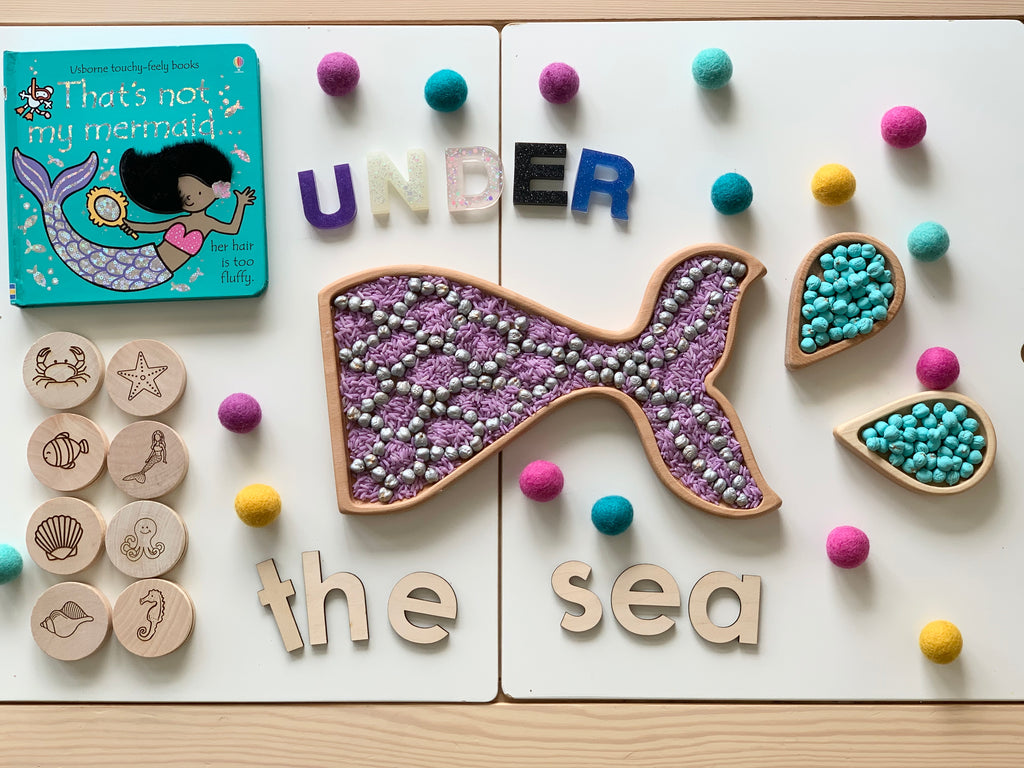 Wooden sensory tray shaped like a whale and raindrops filled with colorful beads with letters spelling UNDER the sea and wooden sea creature tokens