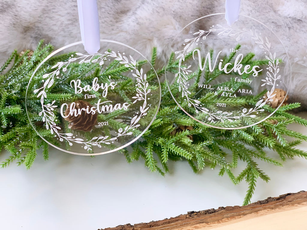 Two engraved clear acrylic Christmas ornaments with white floral wreaths and personalized text hanging over greenery and pinecones