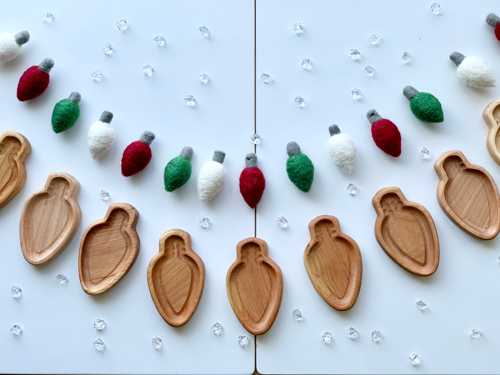 Light bulb shaped natural wooden sensory trays arranged in a row with red, green, and white felted bulbs on white background