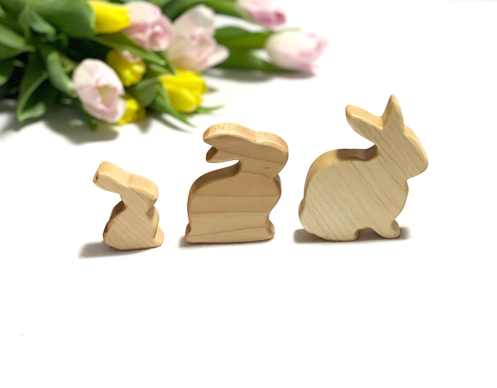 Three smooth wooden toy bunnies of varying sizes arranged in a row with blurred pastel tulips in background