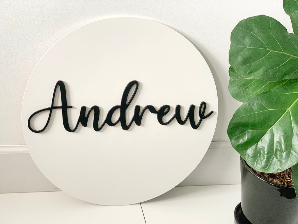 Large white round wooden sign with black cursive 3D name lettering on white floor next to green potted plant