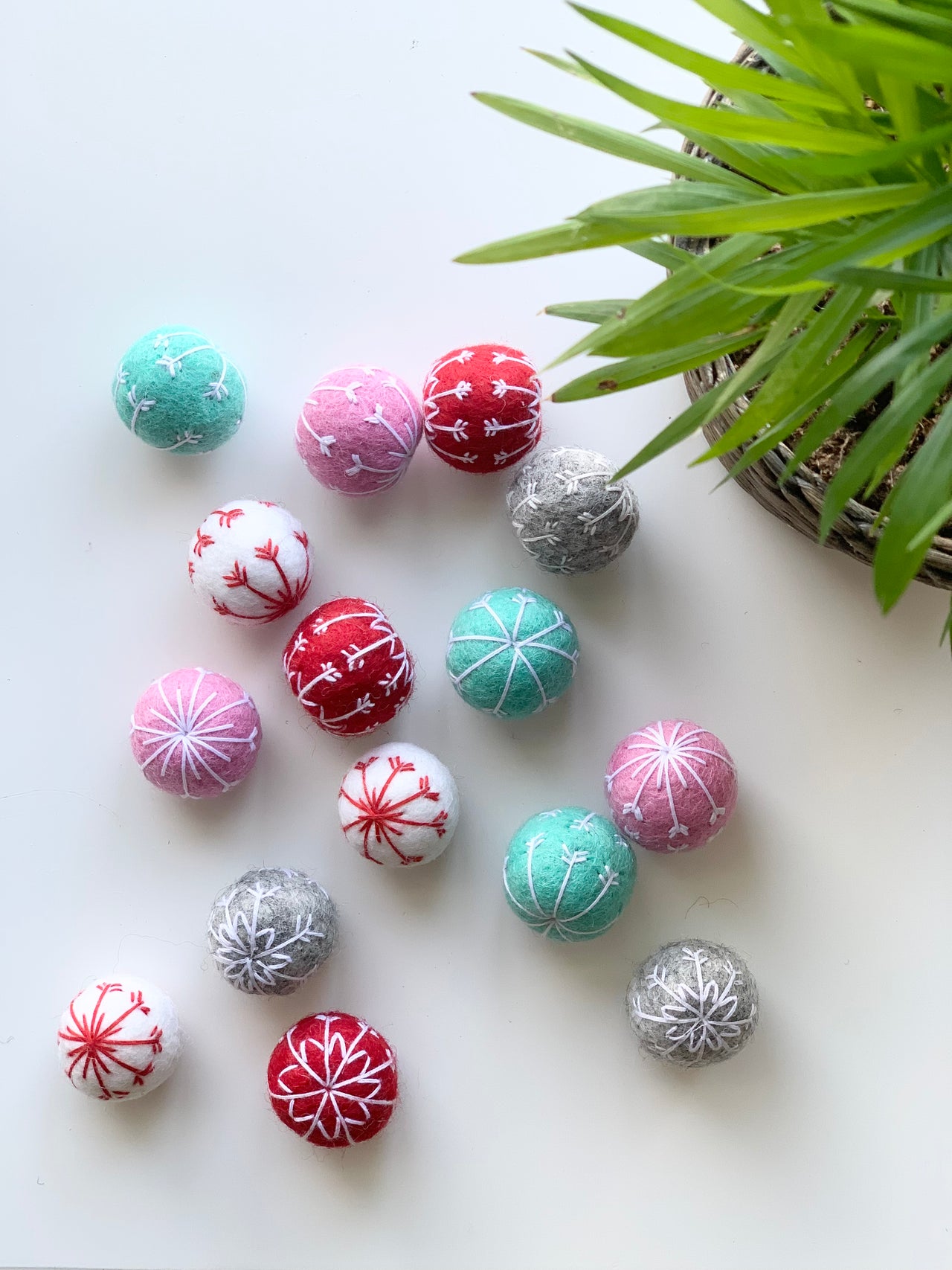 Colorful handmade felt snowflake balls in teal, pink, red, white, and gray arranged on white surface with green plant