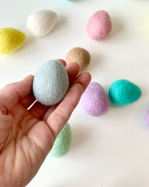 Hand holding one light blue felt Easter egg with other pastel-colored felt eggs on white surface