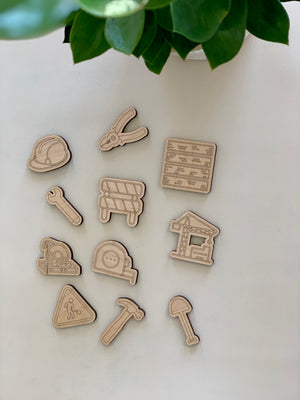 Set of 11 wooden construction and worksite themed loose parts including helmet, tools, caution sign, and building materials on white surface