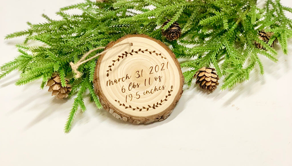 Wood burned wooden baby’s first Christmas ornament with birth details surrounded by pine branches and pinecones