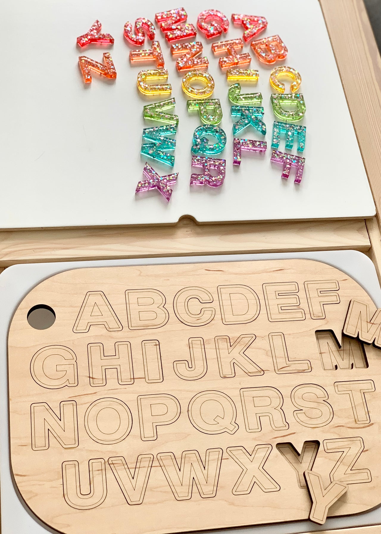 Wooden uppercase alphabet puzzle board with colorful resin letters laid out above on white surface and wood table edge