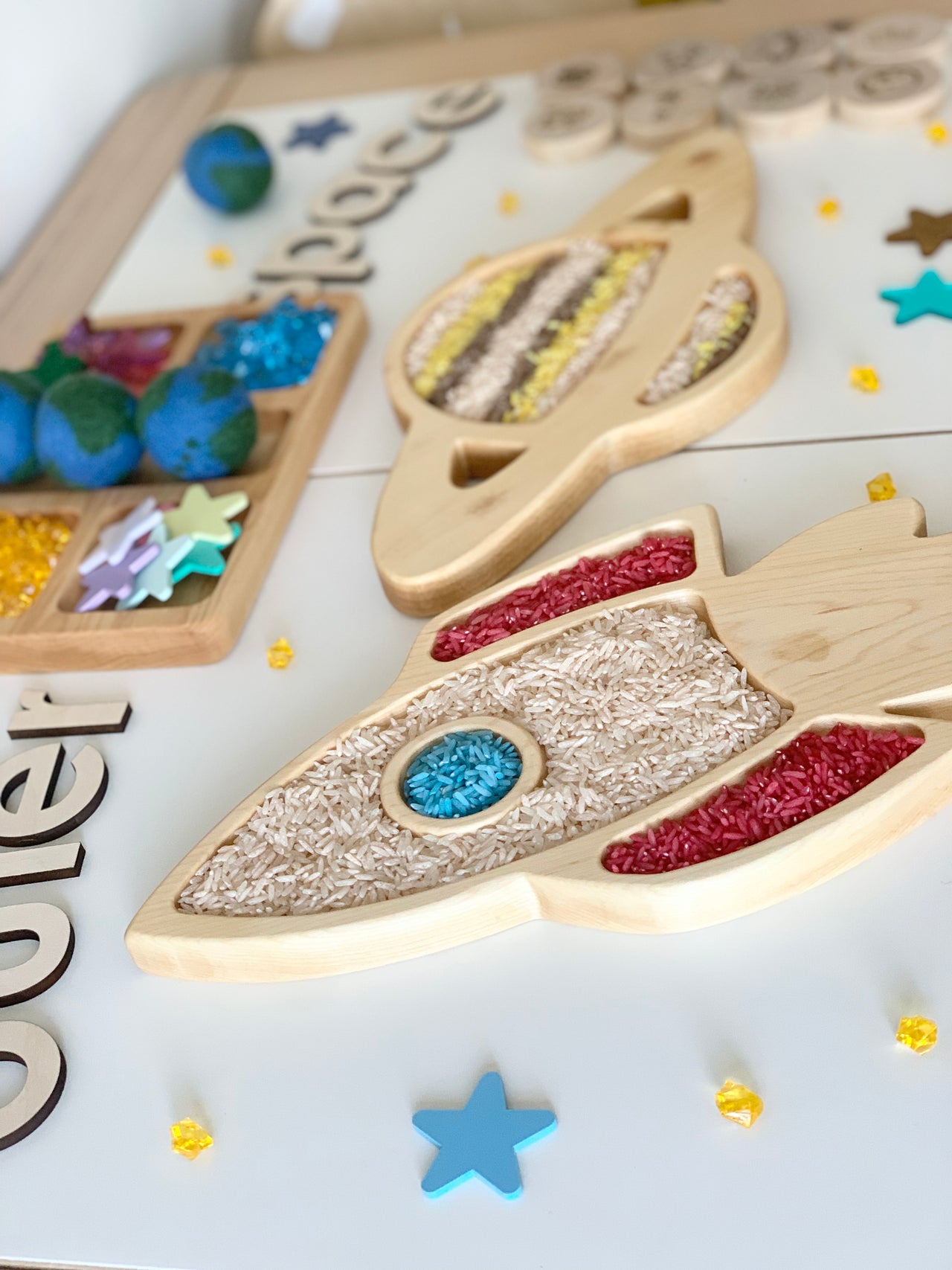 Wooden sensory trays shaped like Saturn and a rocket filled with colored rice on white surface with stars