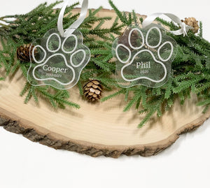 Two clear acrylic paw print Christmas ornaments engraved with pet names and dates on wood slice with pine branches