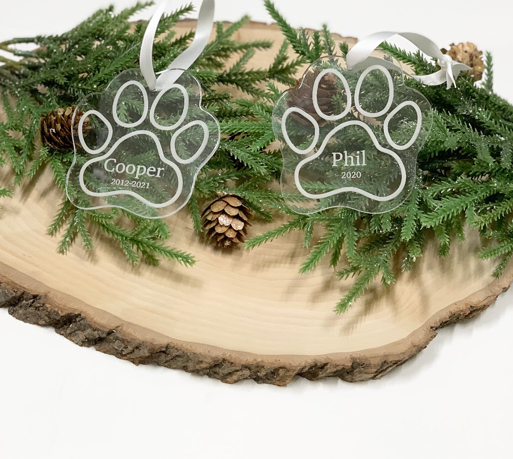 Two clear acrylic paw print Christmas ornaments engraved with pet names and dates on wood slice with pine branches