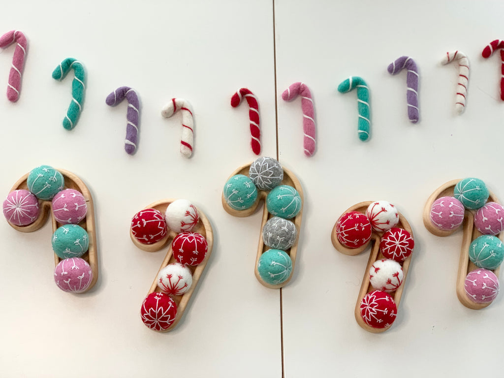 Natural wooden sensory trays shaped like candy canes filled with red, white, teal, gray, and pink soft balls on white surface