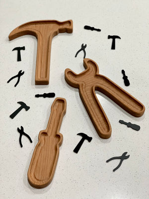 Wooden sensory tray shaped like hammer, pliers, and screwdriver with black tool-shaped cutouts on white surface