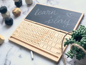 Wooden laptop toy with engraved keyboard and chalkboard screen displaying handwritten learn plus play on marble background