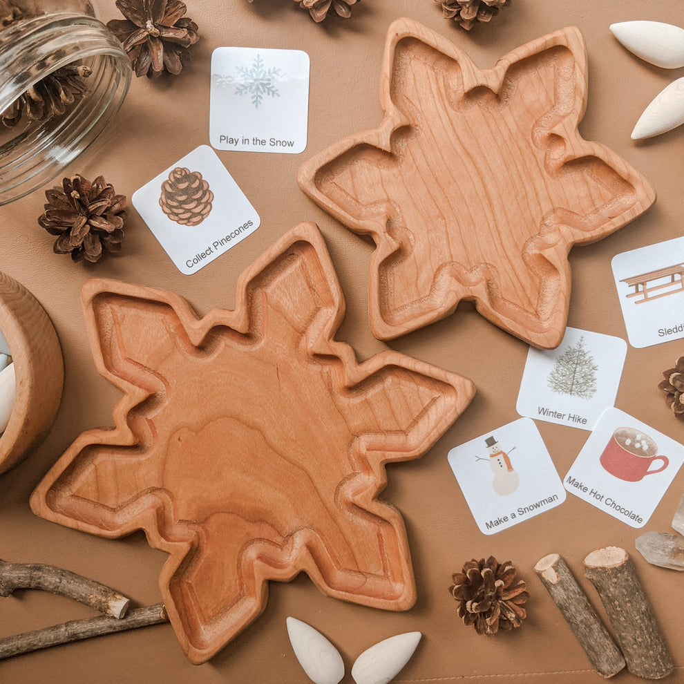 Two natural wooden snowflake-shaped small plates with winter-themed activity cards and pinecones on brown surface