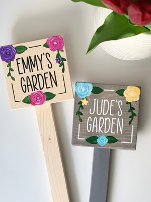 Two small square wooden garden signs on stakes painted with names and colorful floral decorations on wood backgrounds