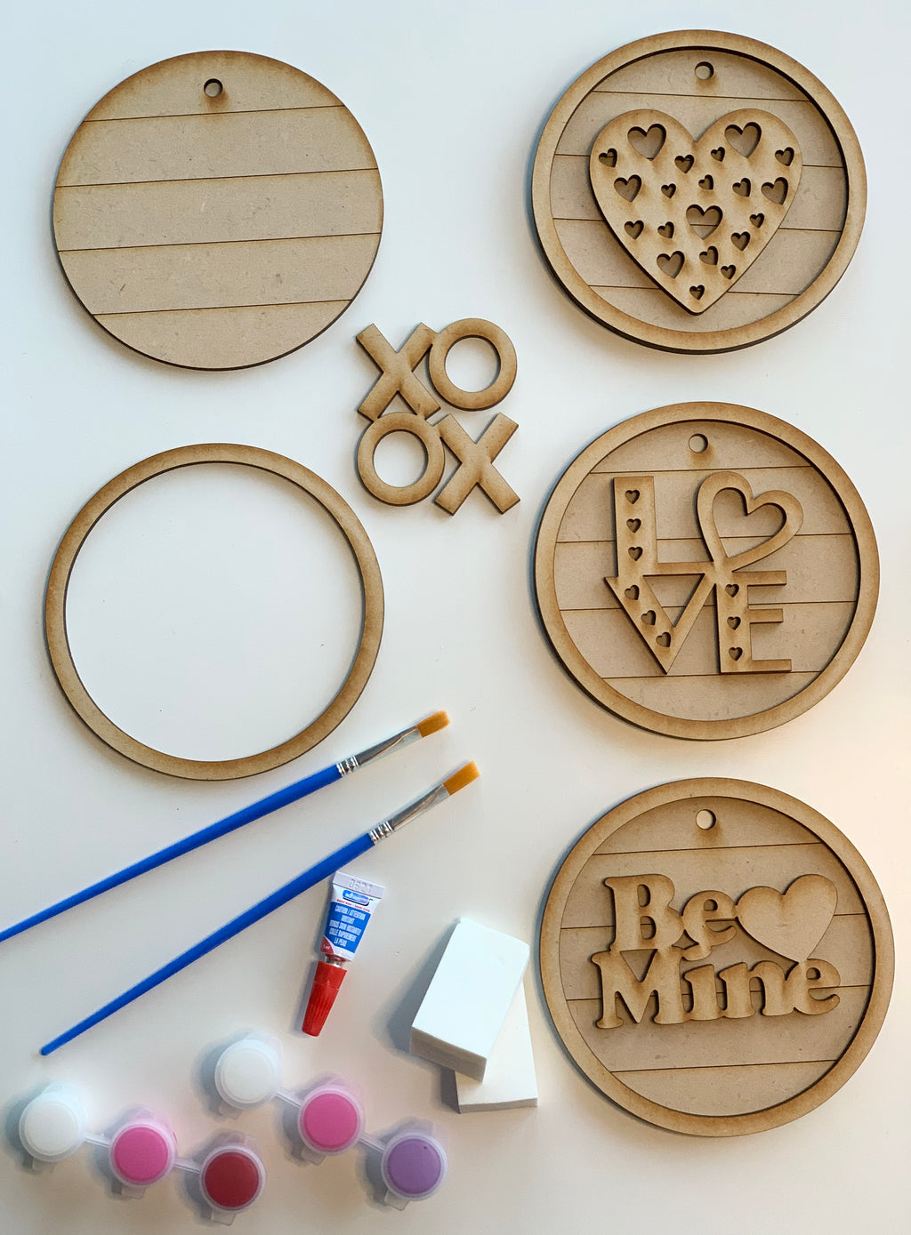 Unpainted wooden love-themed DIY kit with four hanging signs, paint pots, brushes, and sponge applicators on white background