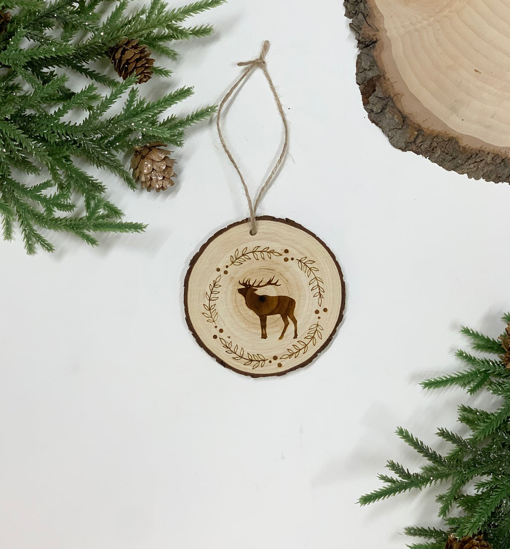 Round wood Christmas ornament with wood burned deer and decorative wreath design hanging with twine on white background
