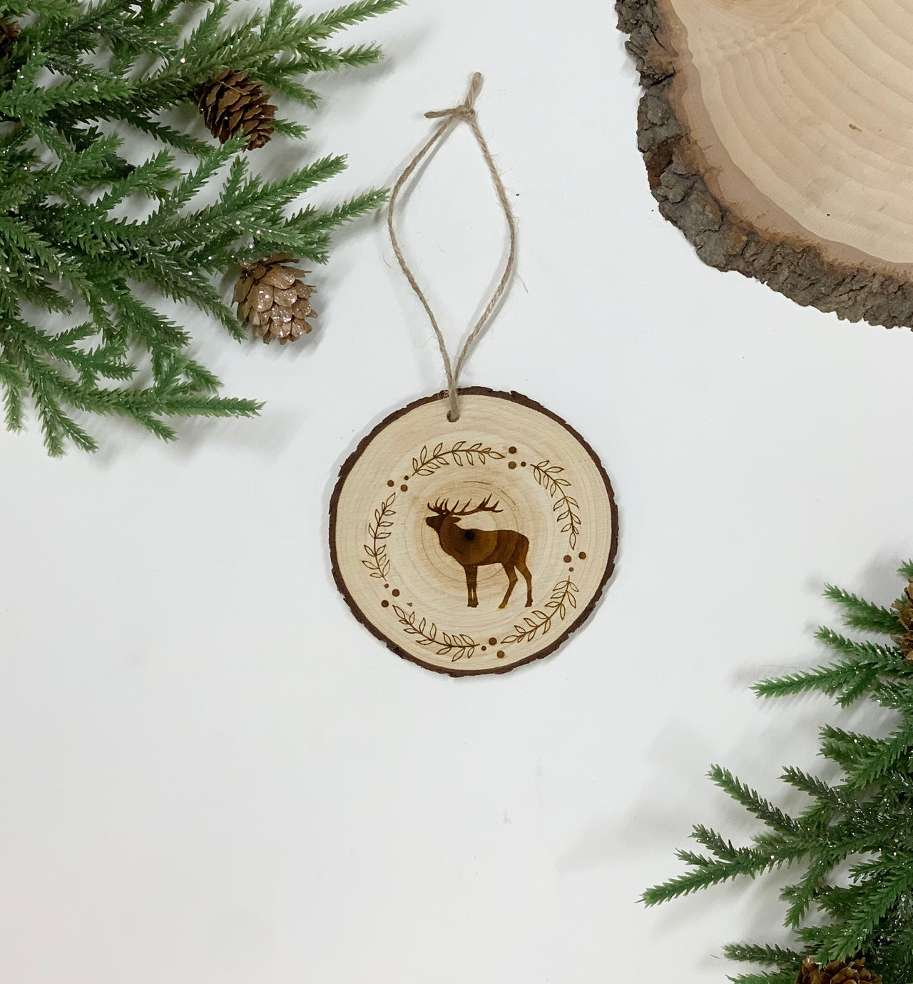 Round wood Christmas ornament with wood burned deer and decorative wreath design hanging with twine on white background