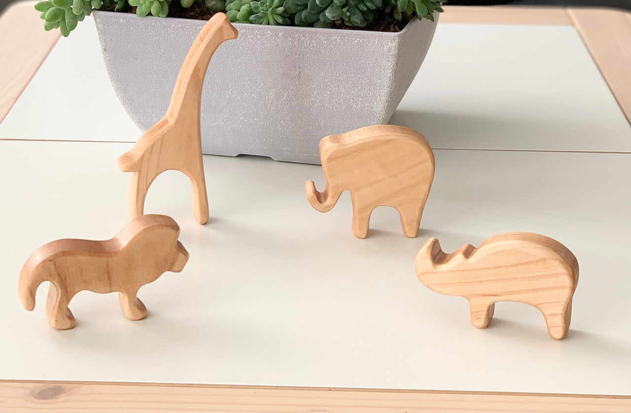 Natural unfinished wooden safari animal figurines including giraffe, elephant, lion, and rhinoceros on white surface