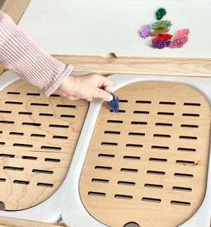 Hand placing blue glittery shape into Flisat table top insert slot with colorful play pieces on light wood surface