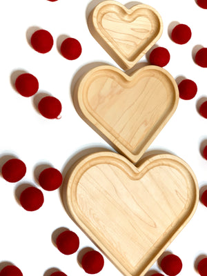 Three natural wooden nesting heart plates with smooth finish surrounded by scattered red felt balls on white background